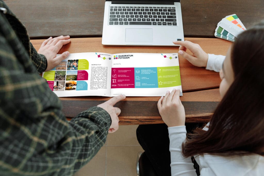 Zwei Personen betrachten gemeinsam eine offene Broschüre am Holztisch mit Laptop.
