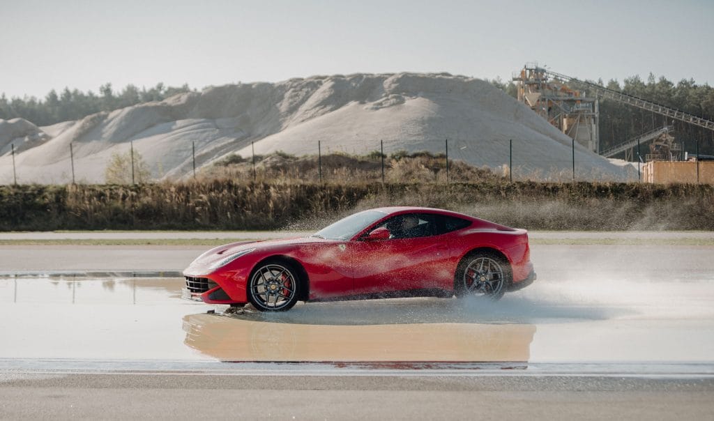 Roter Sportwagen fährt durch Wasser auf Straße mit sandiger Landschaft im Hintergrund.