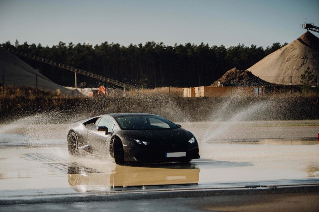 Schwarzer Sportwagen fährt durch Wasserfontäne auf nasser Straße bei sonnigem Wetter.