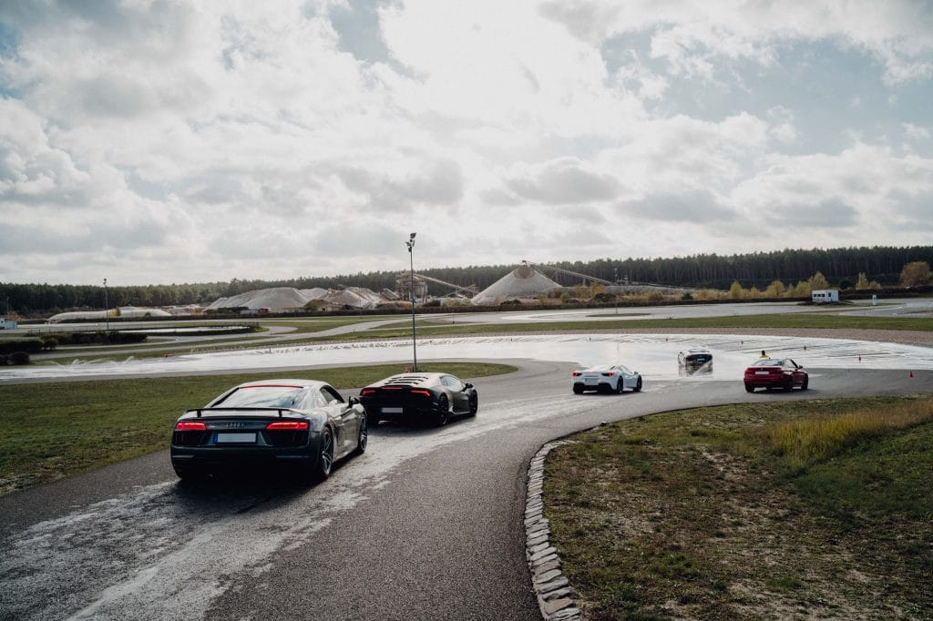 Sportwagen auf Rennstrecke bei nassen Bedingungen, umgeben von Natur und Wolken.