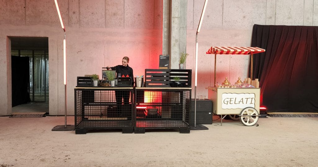Frau hinter Bar mit Beleuchtung, daneben ein Gelati-Wagen in industriellem Ambiente.