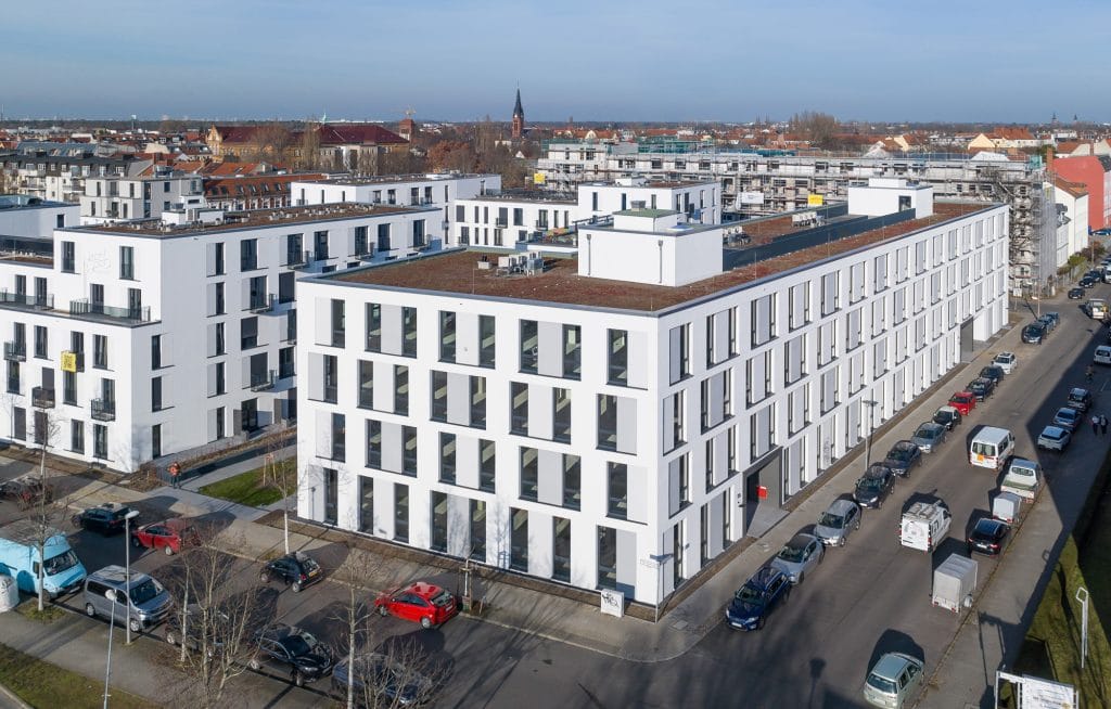 Moderne Wohngebäude in städtischer Umgebung mit parkenden Autos und blauem Himmel.