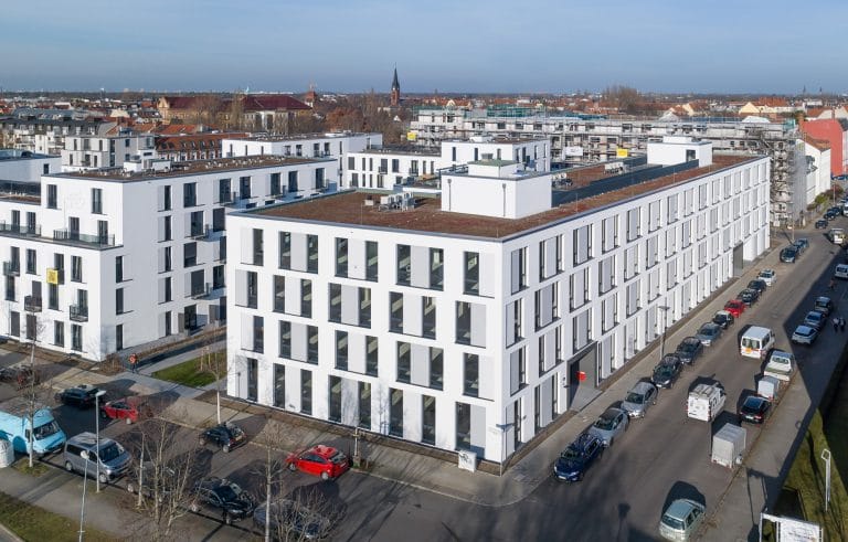 Moderne Wohngebäude in städtischer Umgebung mit parkenden Autos und blauem Himmel.
