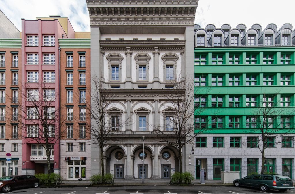 Drei bunte Gebäude nebeneinander in einer städtischen Straßenansicht an einem bewölkten Tag.
