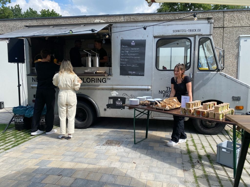 Personen kaufen Essen an einem Foodtruck auf einem gepflasterten Platz bei sonnigem Wetter.