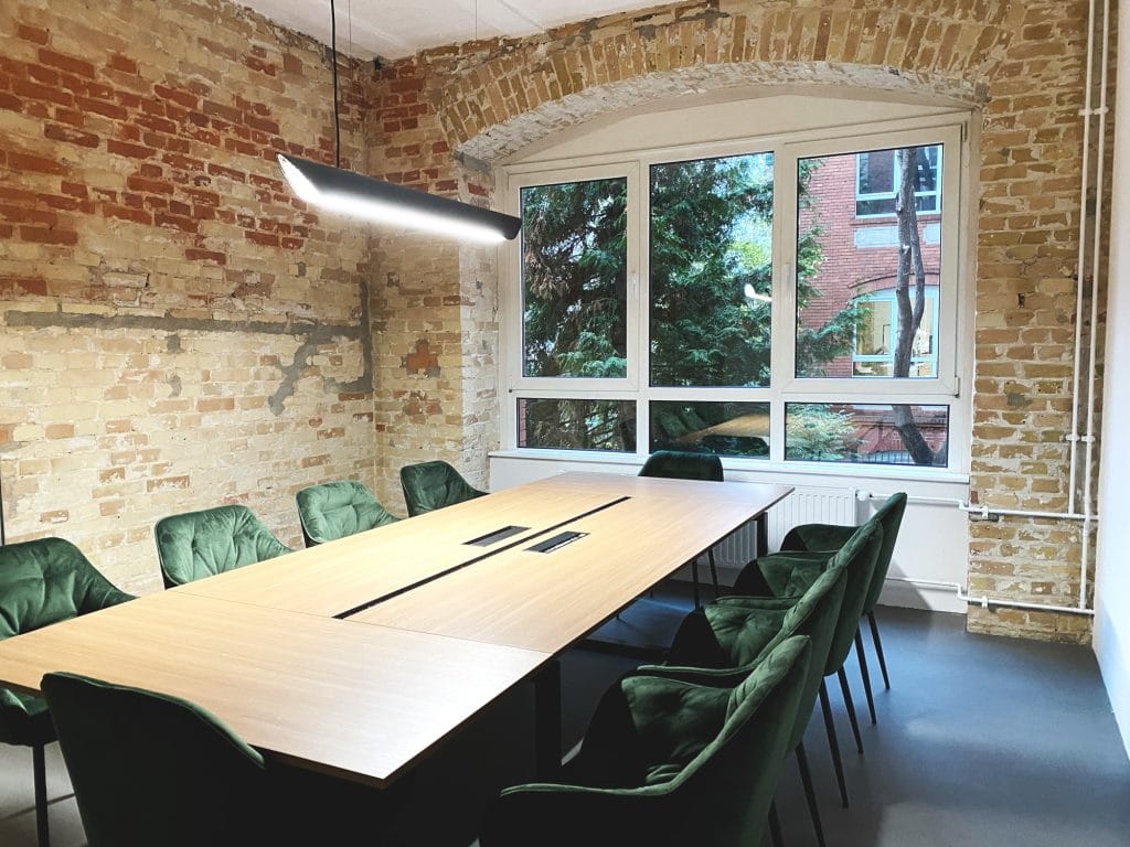 Moderner Besprechungsraum mit Backsteinwand, großem Tisch und grünen Stühlen, viel Tageslicht.