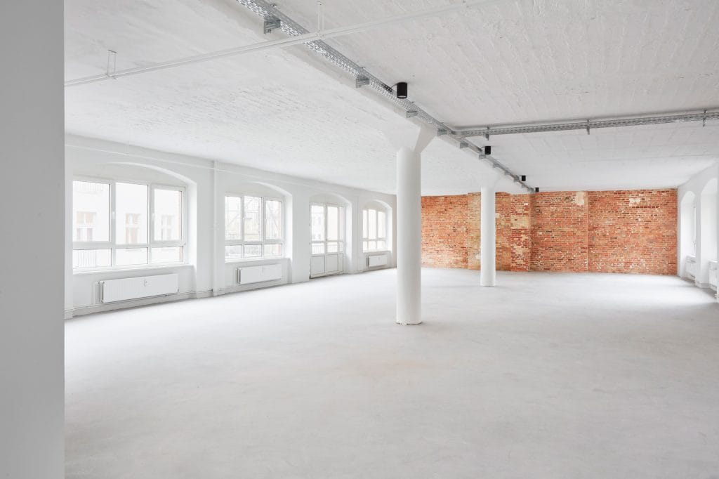 Leerer, lichtdurchfluteter Industrieraum mit Backsteinwand und weißen Säulen.