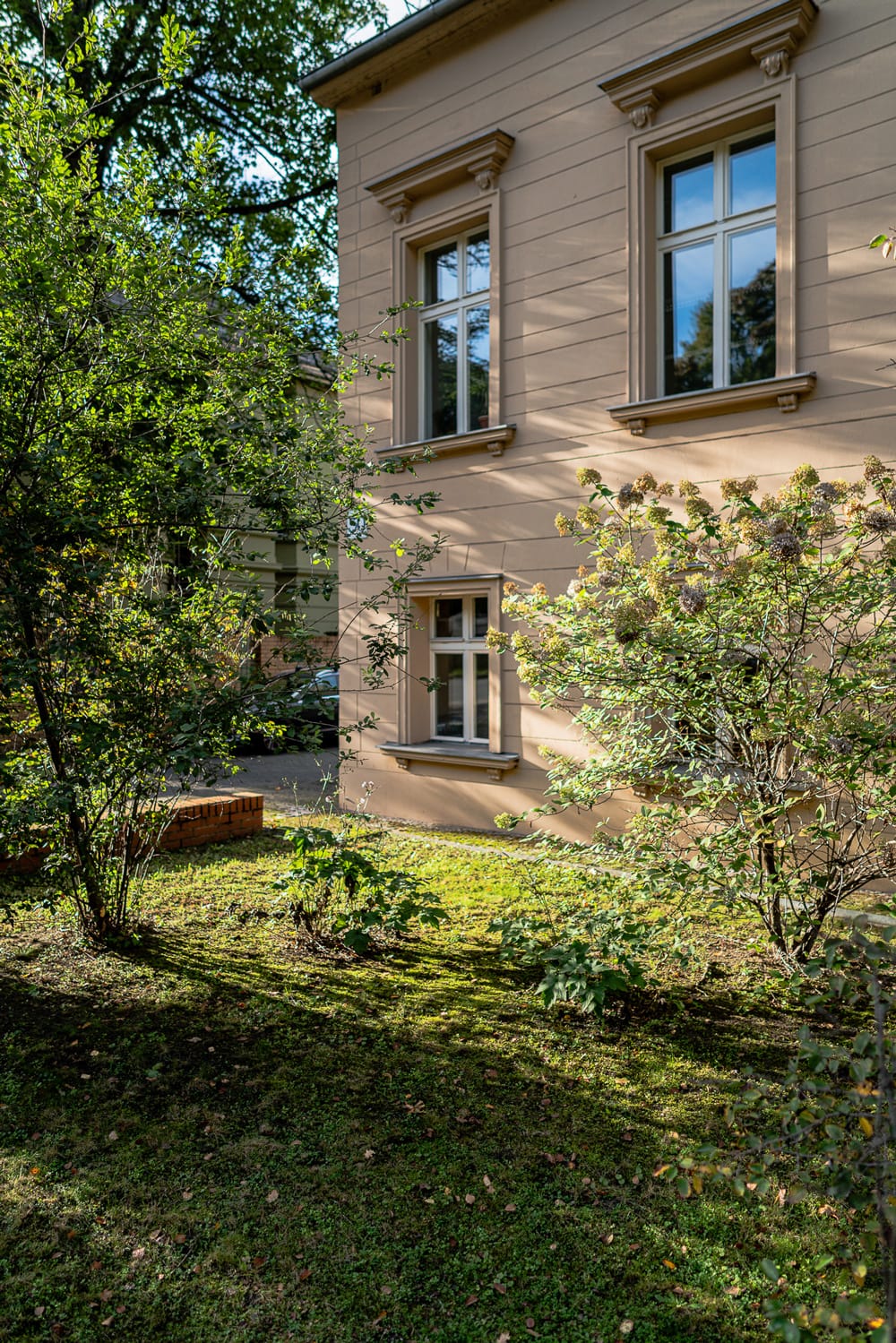 Schattiger Garten eines historischen Hauses mit üppiger Bepflanzung an einem sonnigen Tag.