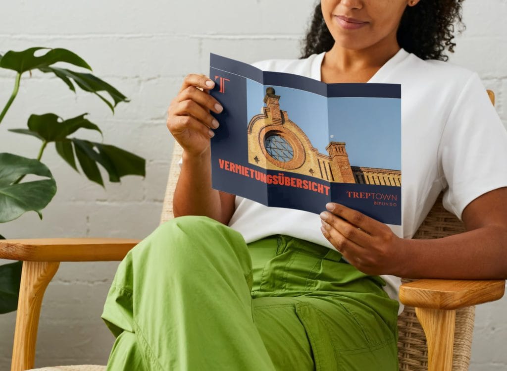 Person liest Broschüre mit Vermietungsübersicht, im Sessel sitzend, neben Pflanze.