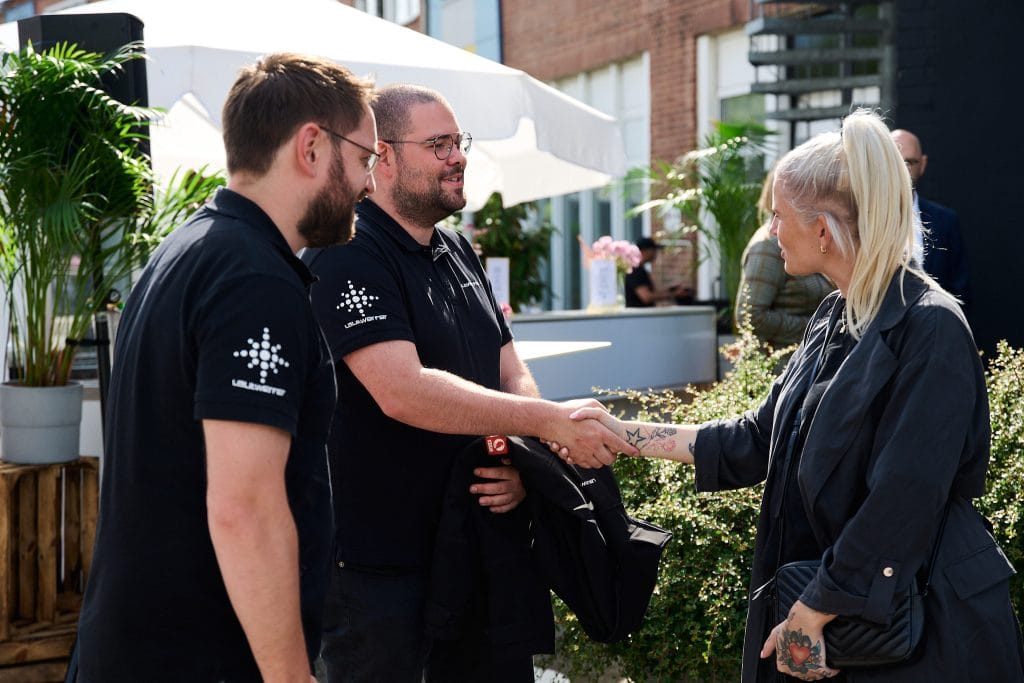 Drei Personen begrüßen sich bei einer Outdoor-Veranstaltung mit Händedruck und Lächeln.
