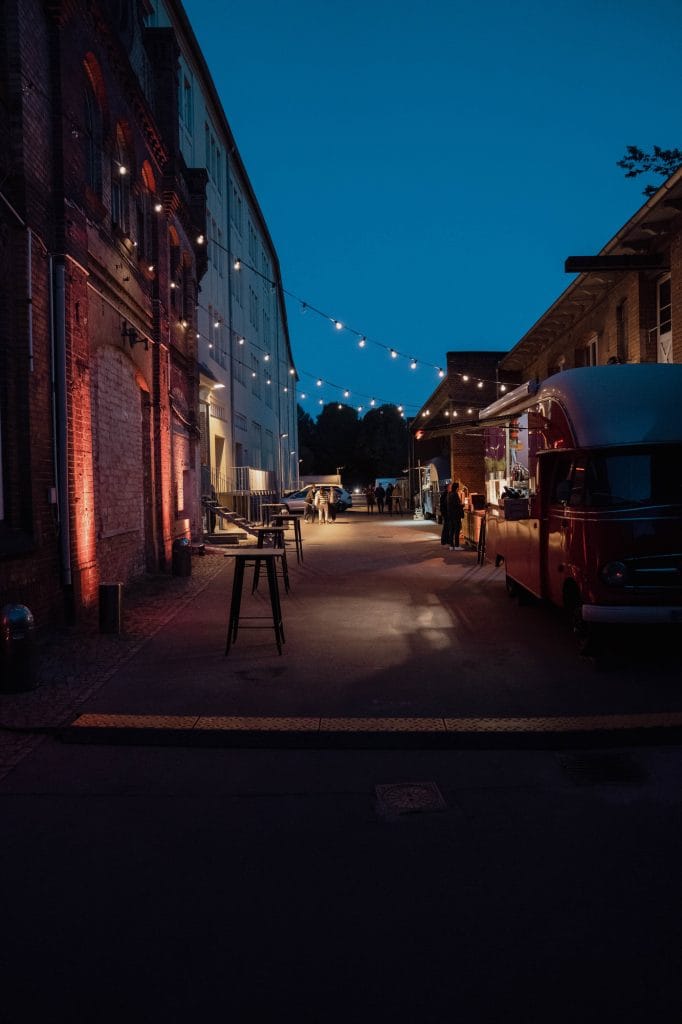 Nachtmarkt mit Lichterketten, rotem Imbisswagen und Industriefassade im Hintergrund.