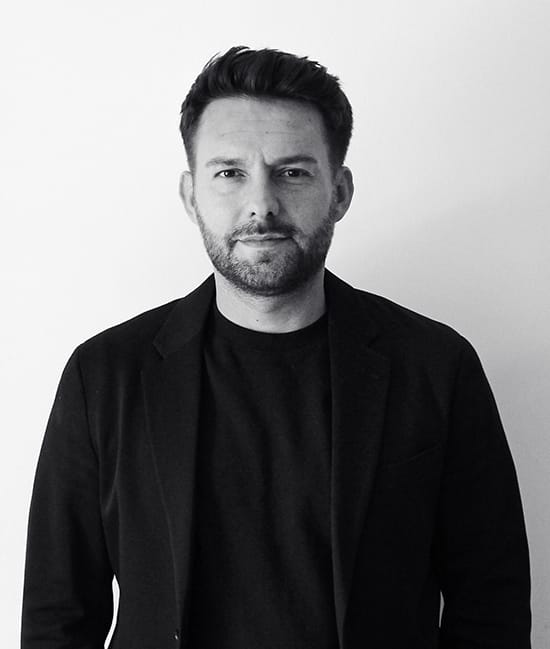 Mann im schwarzen Anzug vor neutralem Hintergrund, freundlicher Ausdruck.