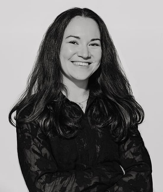 Junge Frau mit langen Haaren und schwarzem Hemd lächelt vor weißem Hintergrund.