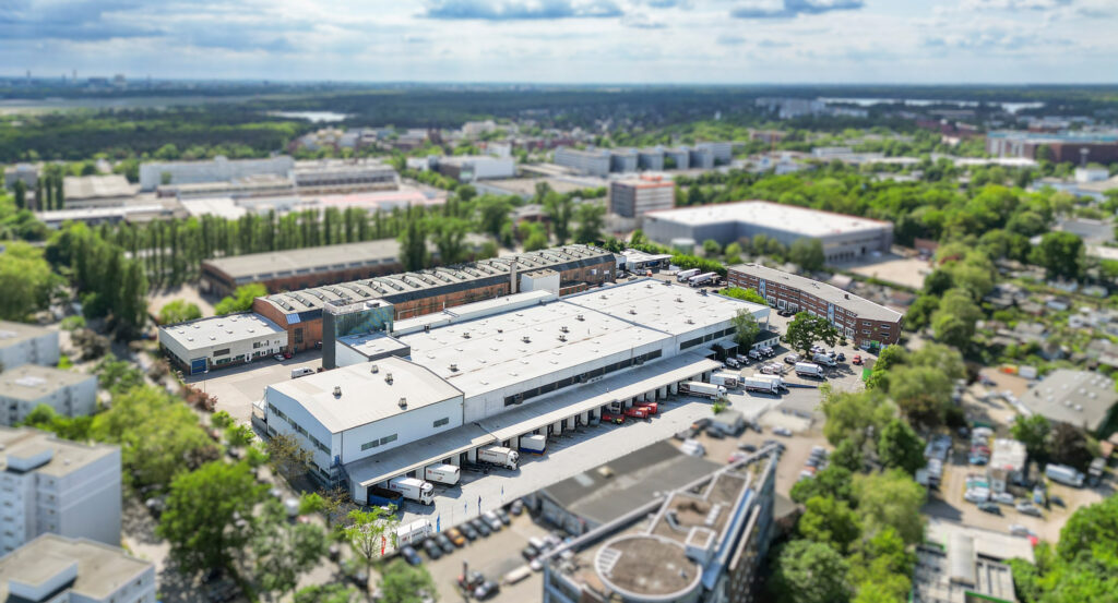 Luftaufnahme eines Logistikzentrums mit umliegenden Gebäuden und grünem Stadtgebiet.